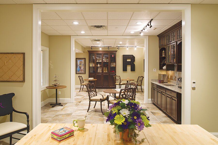 A brightly lit seating area with a table, chairs, and a kitchenette, visible through a large doorway from a front room.