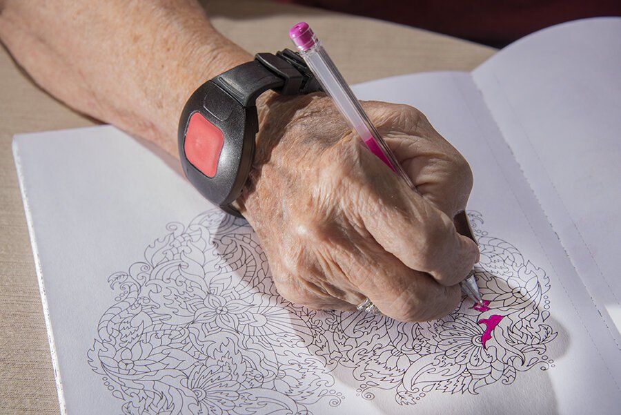 A hand wearing a medical alert bracelet colors a complex floral pattern in a coloring book using a pink marker.