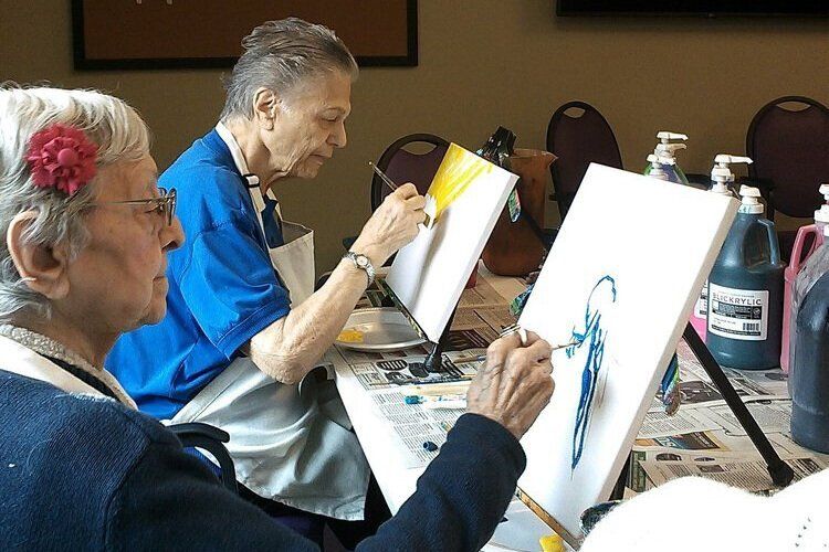Two people painting on canvases at a table in a bright room.