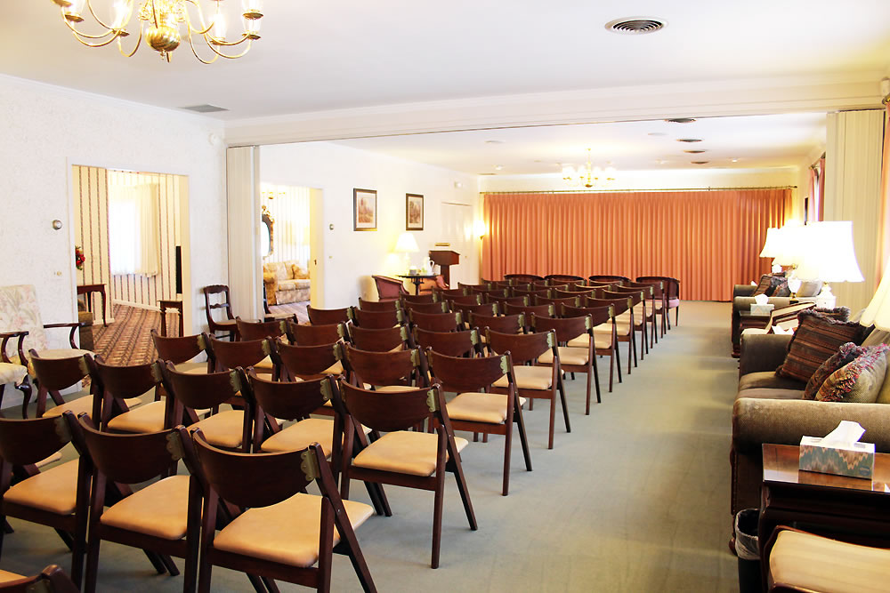 Rows of empty wooden chairs in a funeral home chapel, with sofas and stage with curtain in the background.