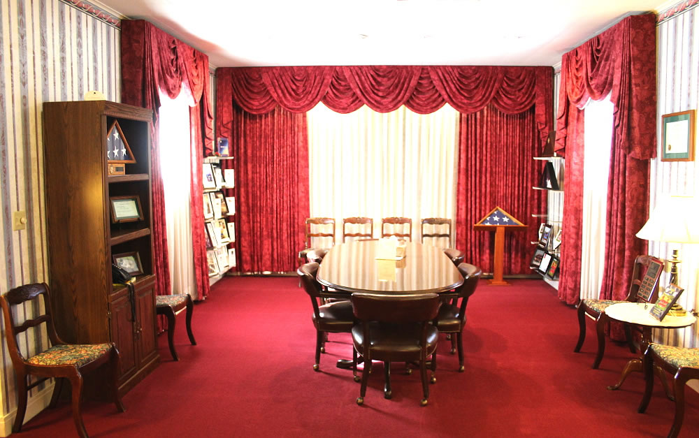 Formal room with red carpet, drapes, and furniture. A large oval table and chairs are in the center.