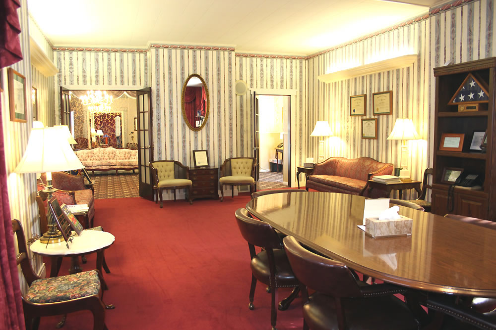 Funeral home viewing room with a red carpet, a long table, and multiple seating areas.
