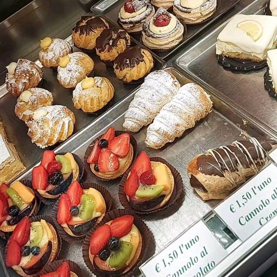 Esposizione di dolci: crostate di frutta, bignè, éclair al cioccolato, croissant e cannoli.