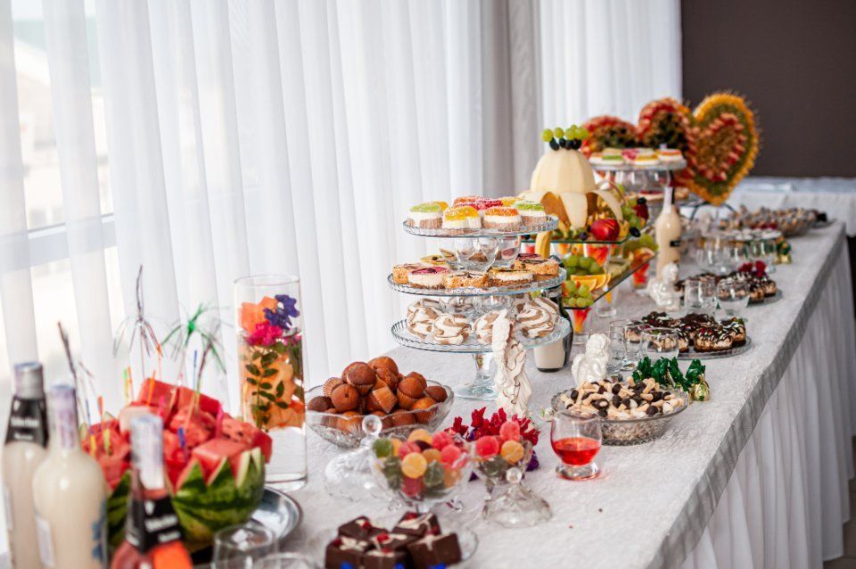 Tavolo del buffet imbandito con dolci, frutta e bevande; tovaglia bianca, sala luminosa.