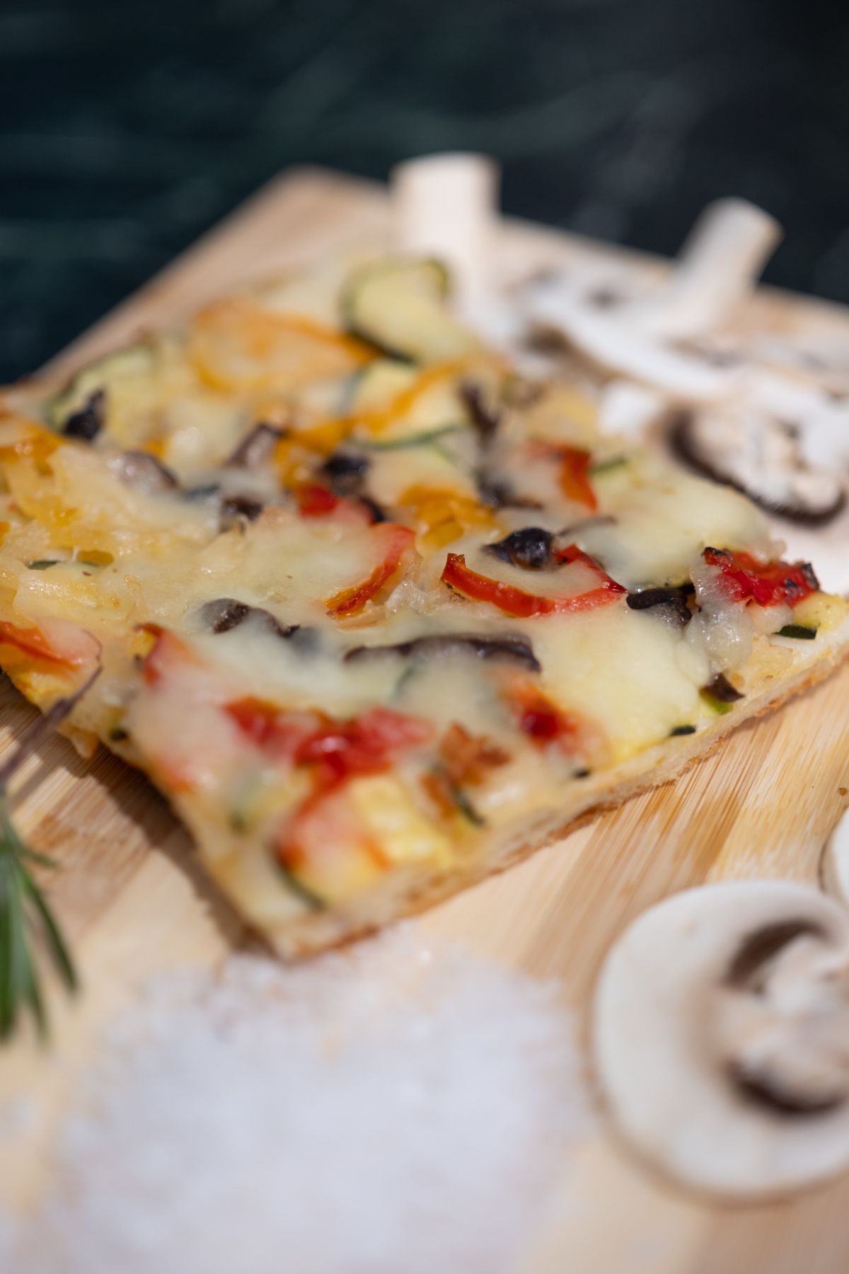 Trancio di pizza con pomodori rossi, zucchine e funghi su un tagliere di legno con sale e altri funghi.