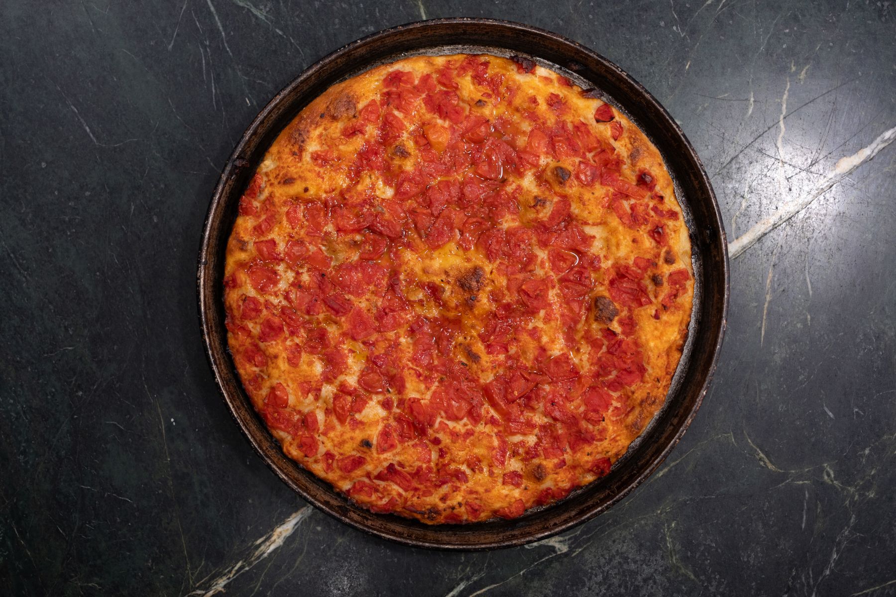 Pizza al pomodoro in una teglia scura su una superficie di marmo nero.