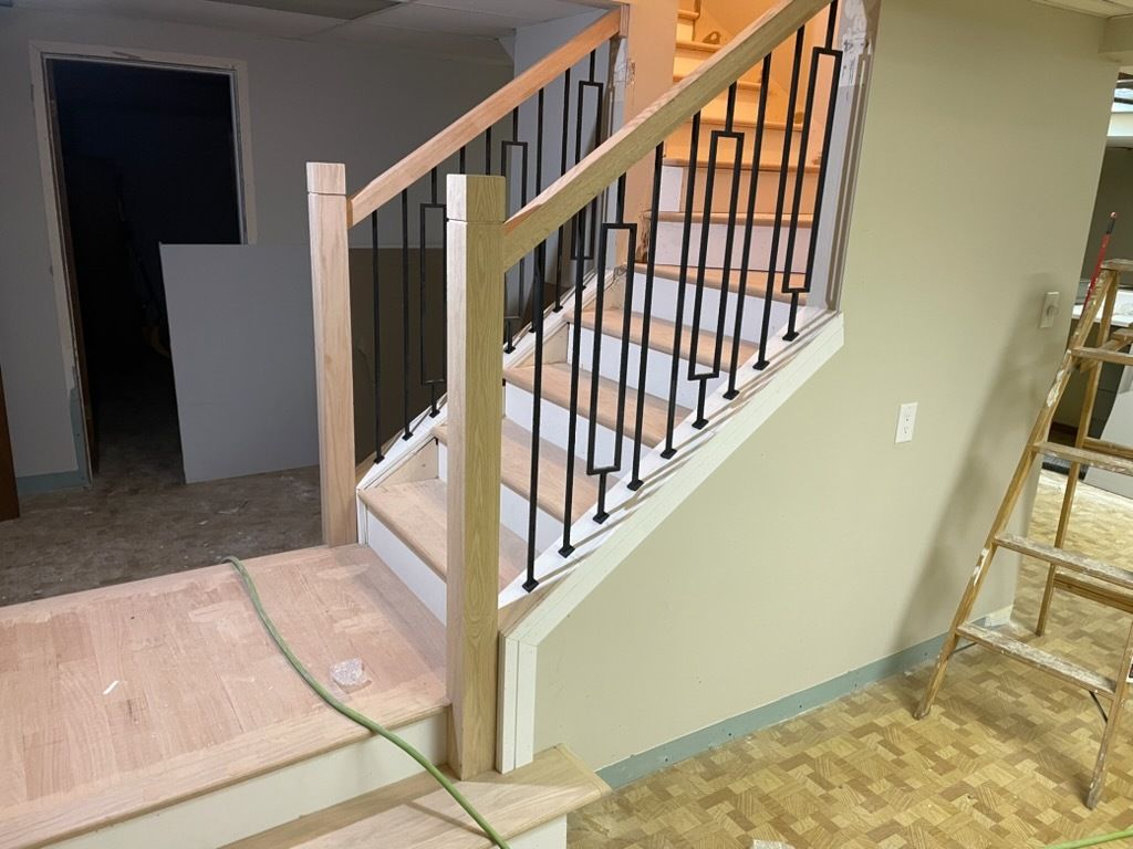 Indoor staircase with wood railing and black balusters beside a beige wall and unfinished floor.
