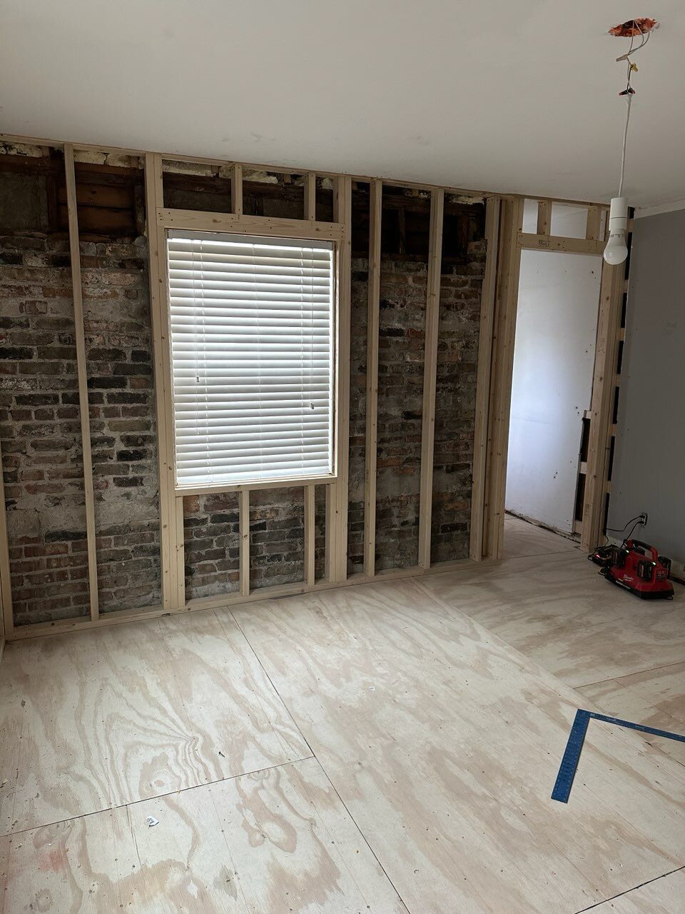 Partially stripped interior wall with exposed studs, window blinds, bare floor, and a doorway under renovation