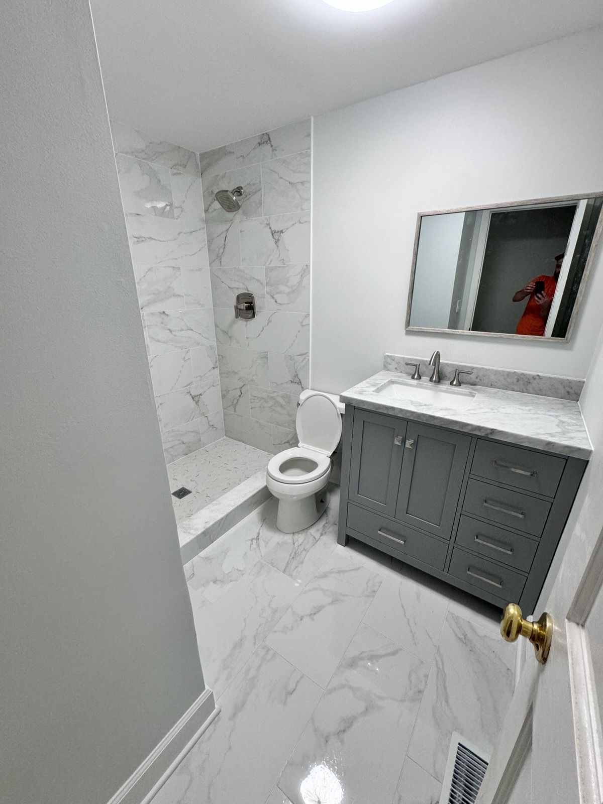 Small white bathroom with marble shower, toilet, gray vanity, and wall mirror.