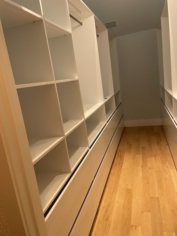 Narrow walk-in closet with white shelves and drawers, beige walls, and light wood floor.