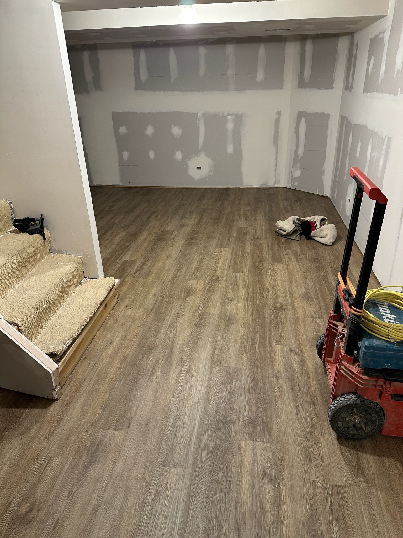 Empty unfinished room with gray drywall, wood floor, sofa at left, and a red dolly at right.