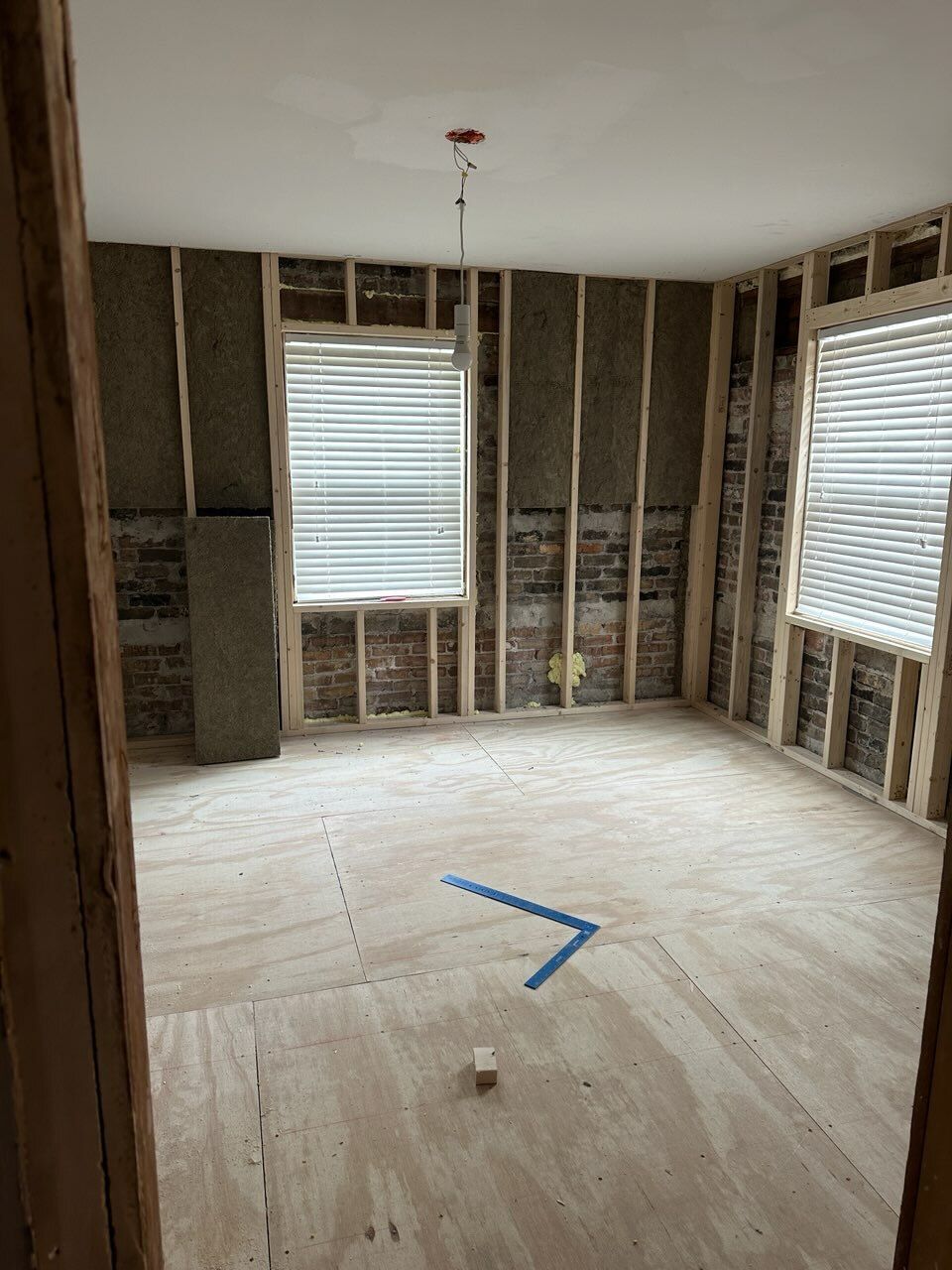 Empty unfinished room with exposed wall studs, two windows with blinds, and bare plywood floor.