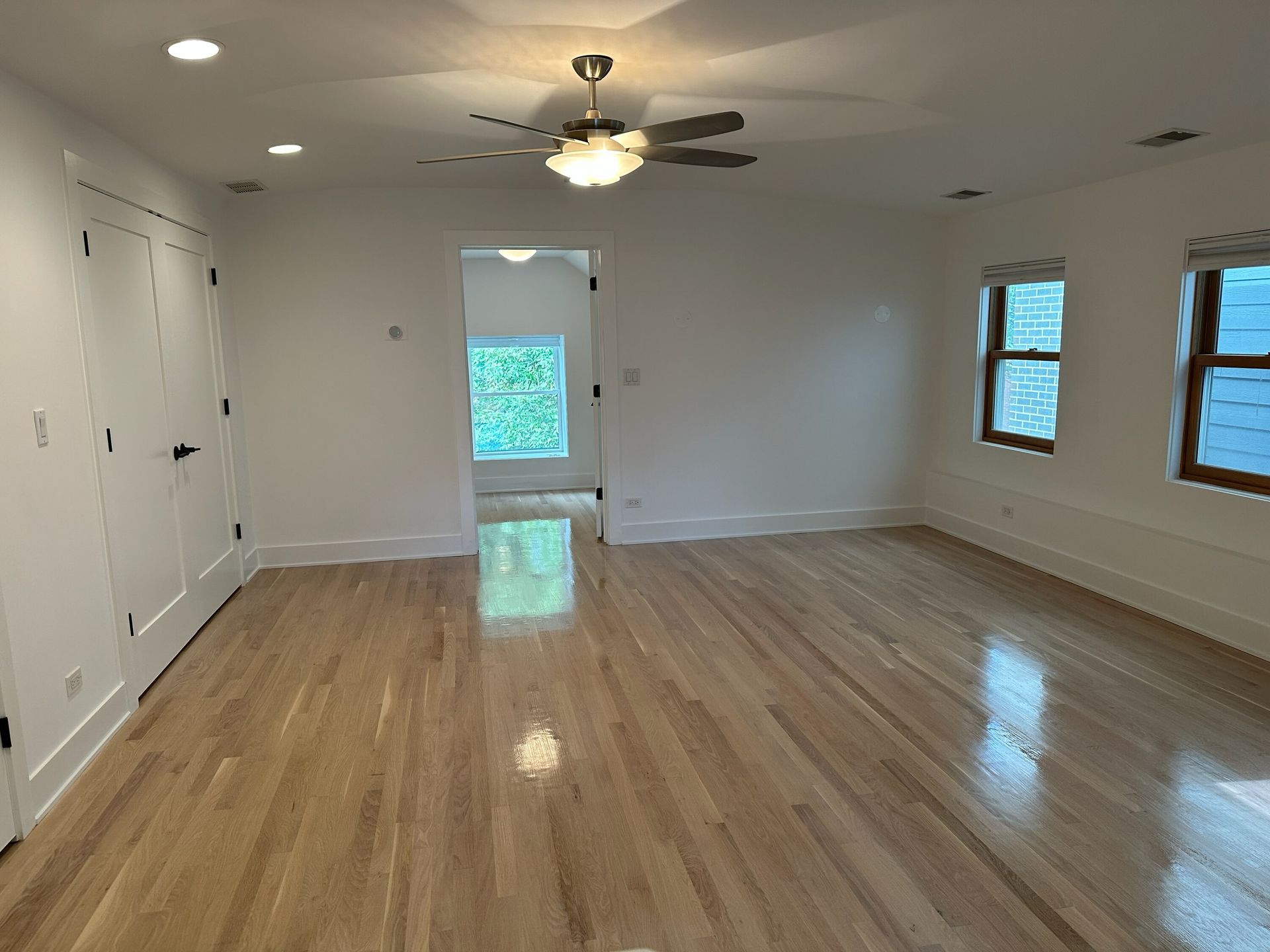 Empty living room with hardwood floors, white walls, ceiling fan, and windows letting in natural light