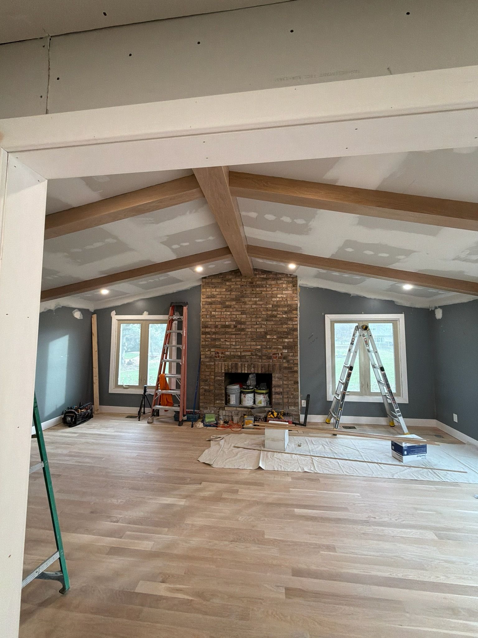 Renovated attic room with exposed beams, brick fireplace, ladders, and tools on a bare wooden floor.