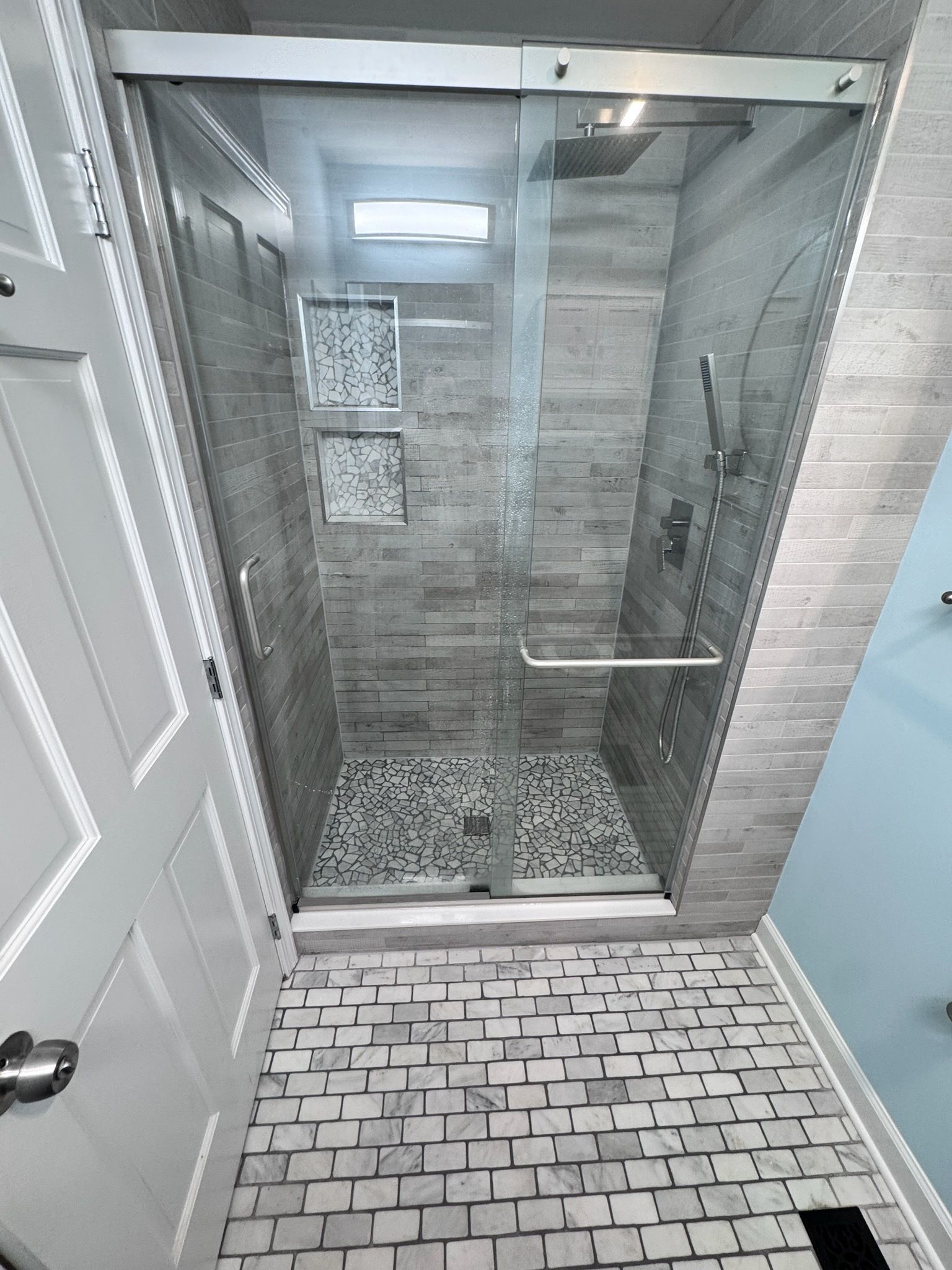 Glass walk-in shower with gray tile walls, pebble floor, and tiled bathroom floor outside.
