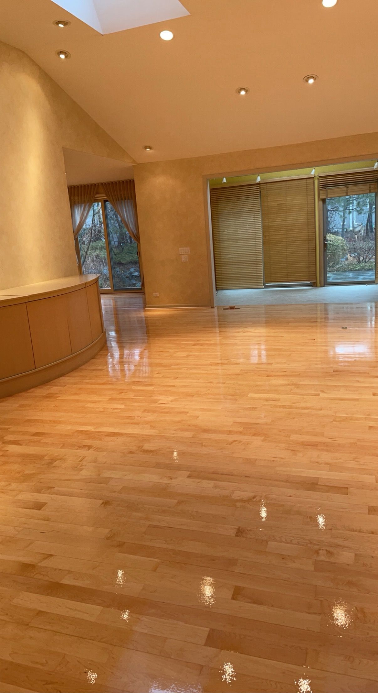 Spacious empty room with polished wooden floor, warm tan walls, recessed lights, and large windows with blinds.