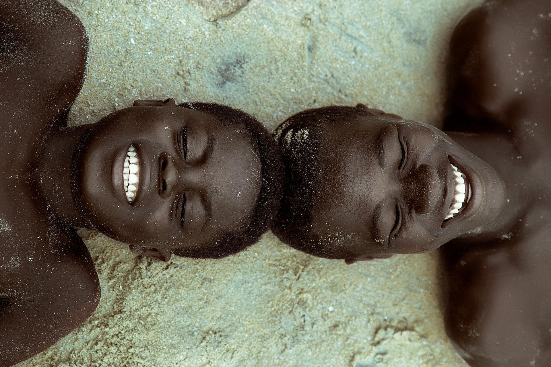 Two boys are laying on their backs in the sand and smiling.in Ghana