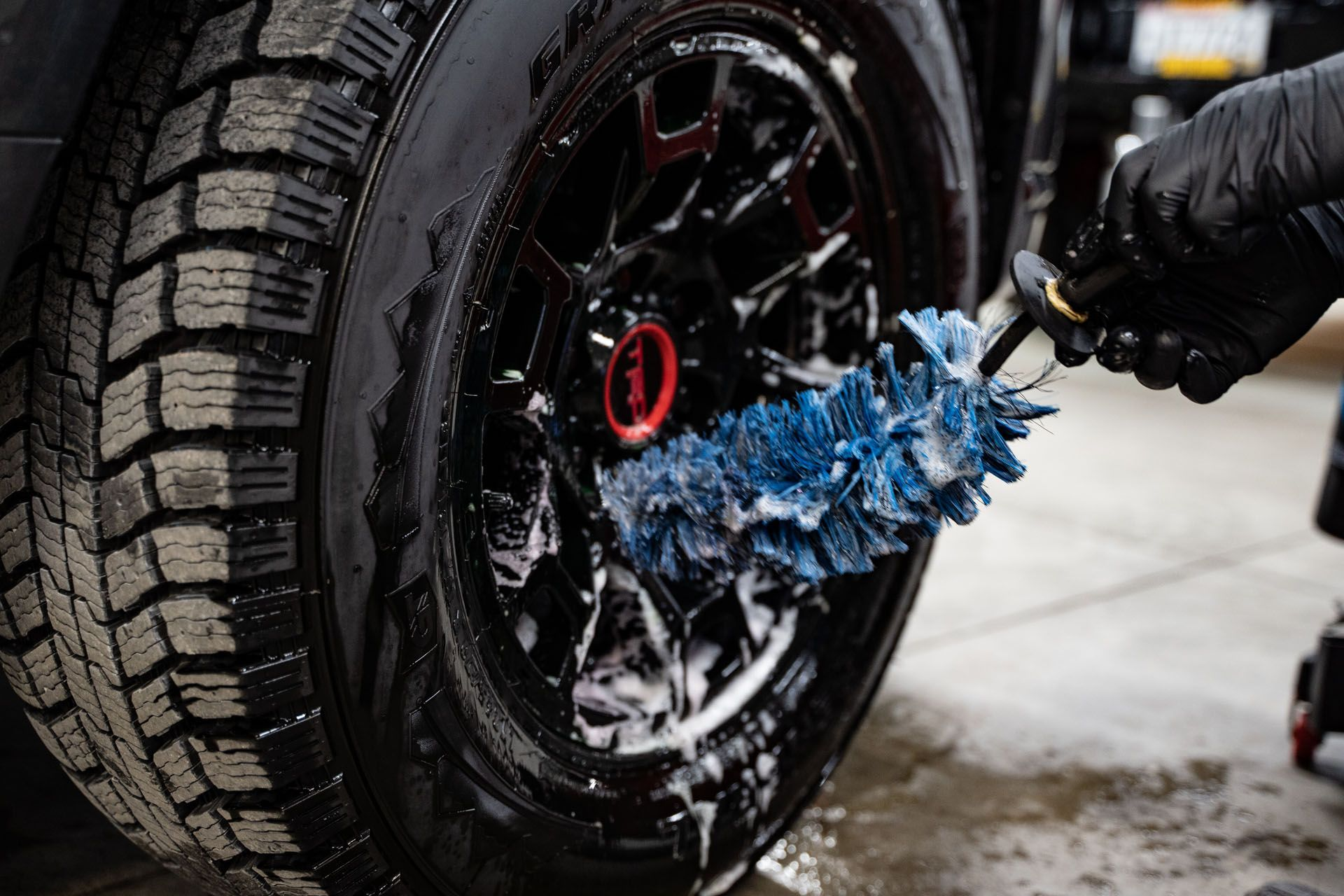 Gloved hand sprays blue water onto a black car tire in a garage.