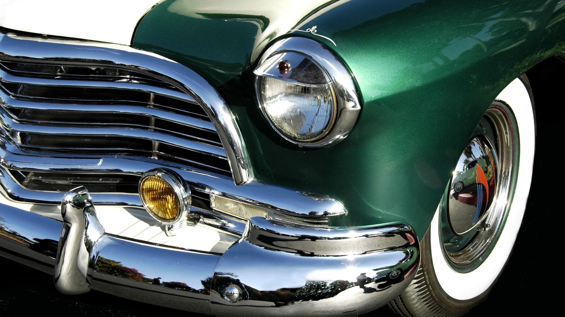 Close-up of a vintage green car’s chrome grille, headlight, and whitewall tire.