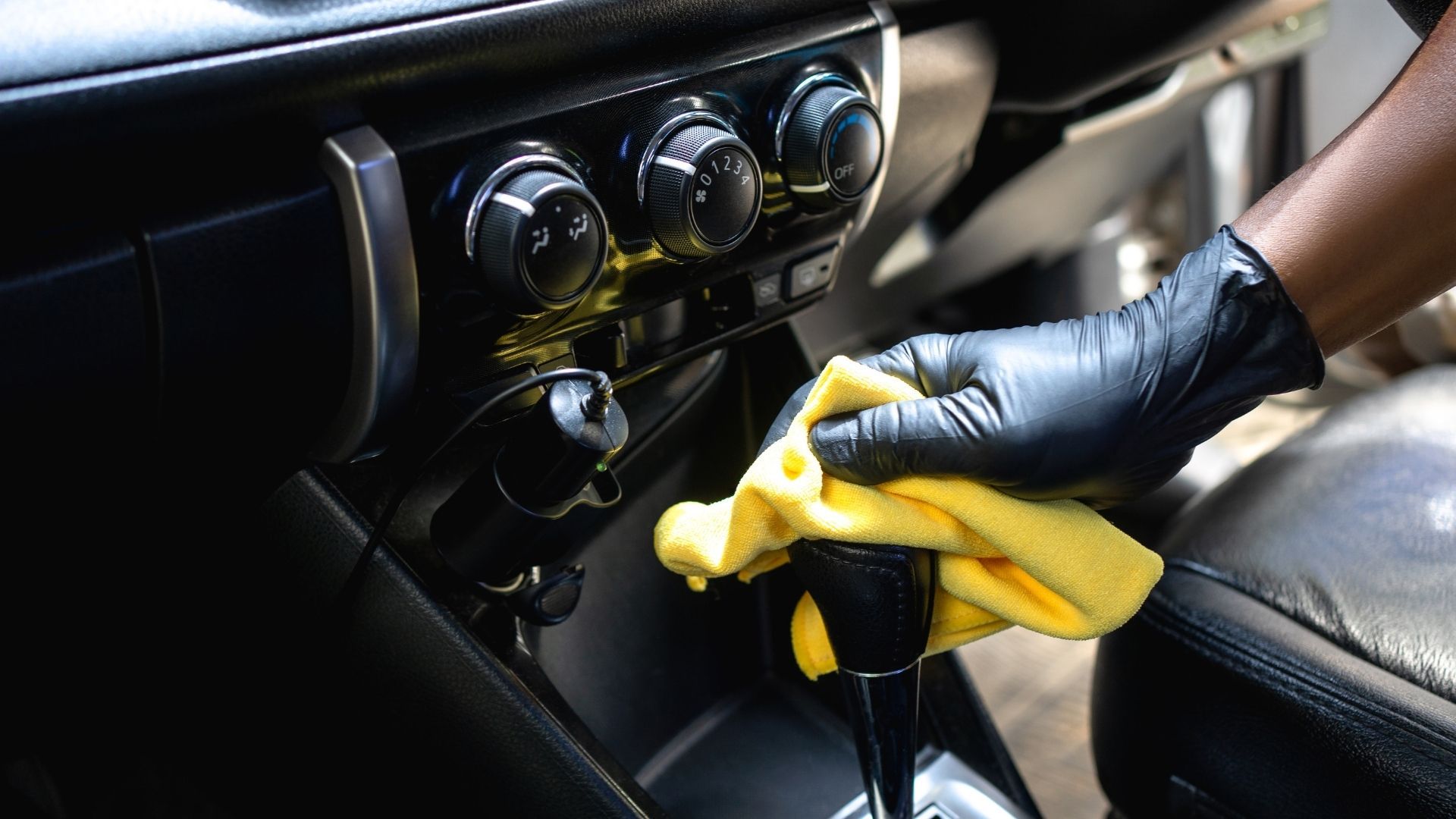 Gloved hand cleaning a car’s center console with a yellow cloth