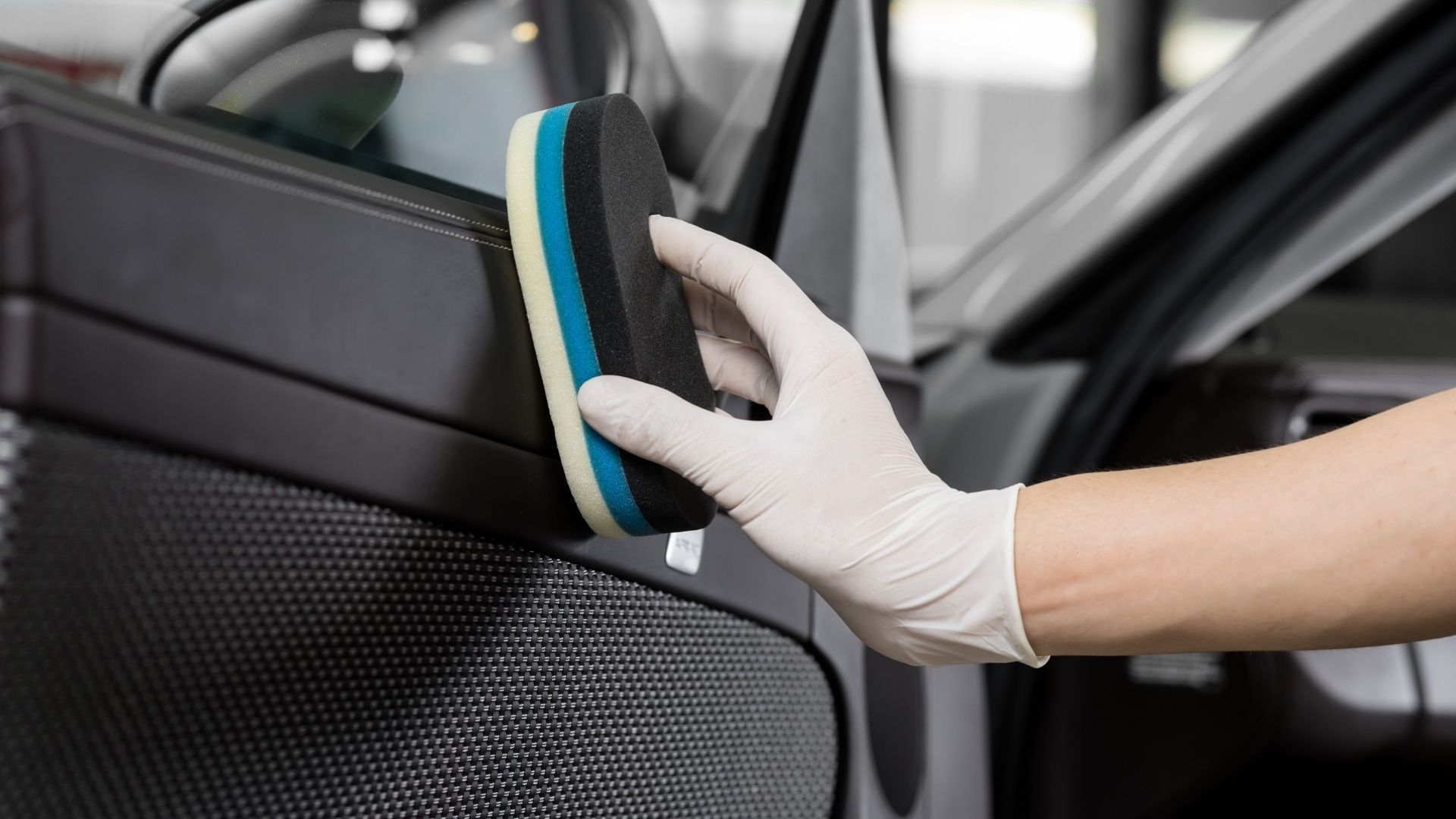 Gloved hand cleaning a car door interior with a black and blue sponge