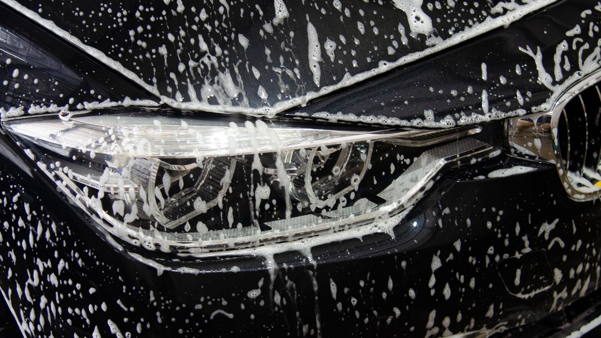 Black car headlight close-up covered in water droplets and soap foam.