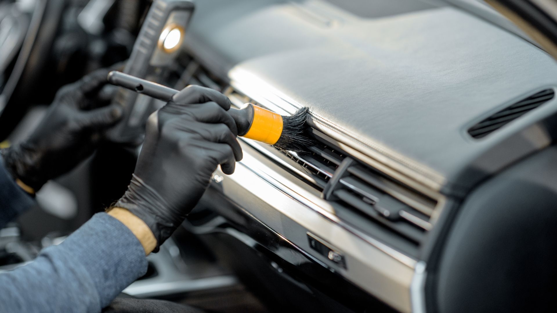 Mechanic cleaning a car dashboard air vent with a brush and cloth gloves
