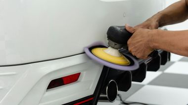 Person polishing a white car bumper with a yellow buffer pad and black cloth