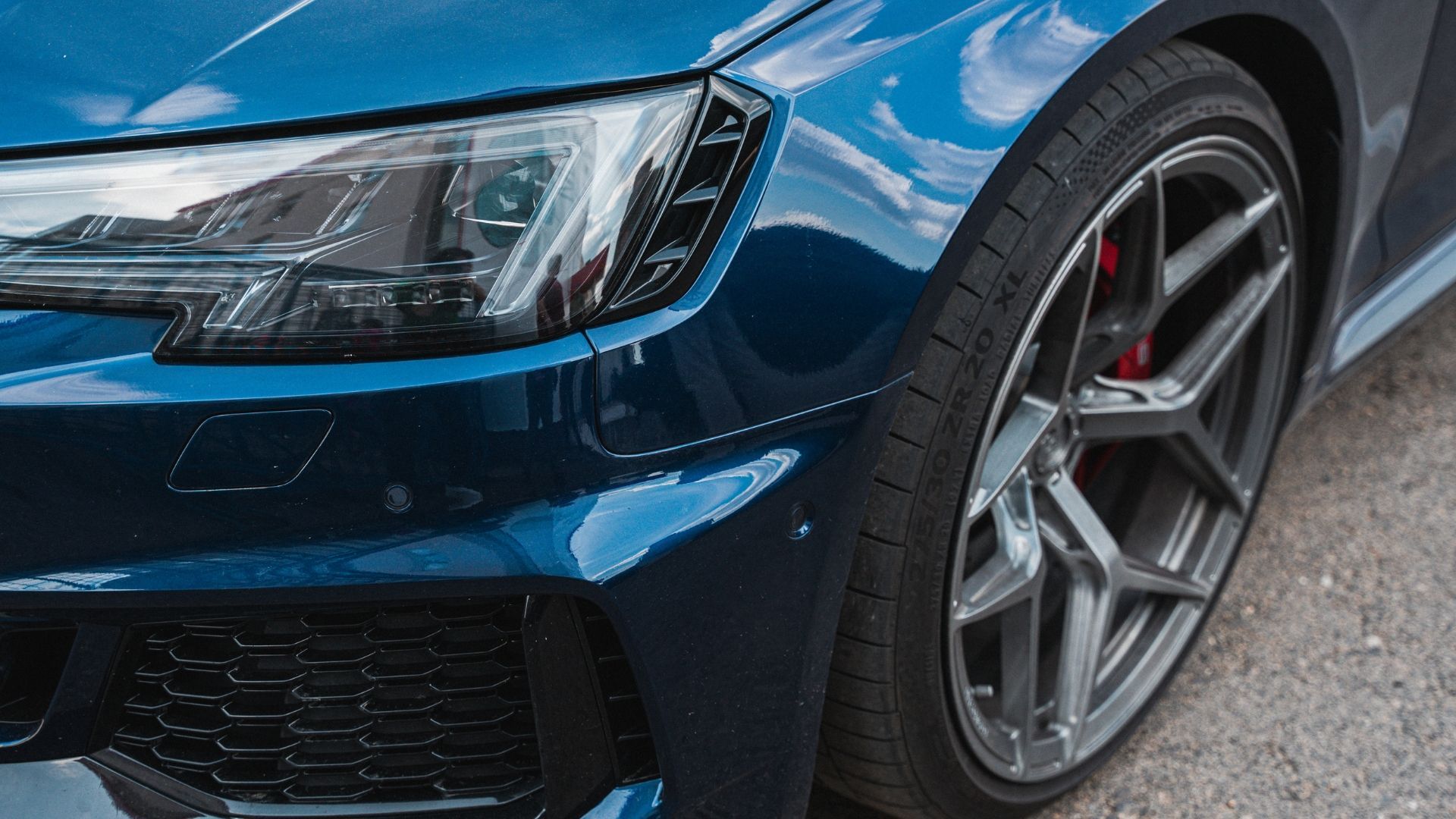 Close-up of a blue sports car’s front wheel, headlight, and grille on a paved road