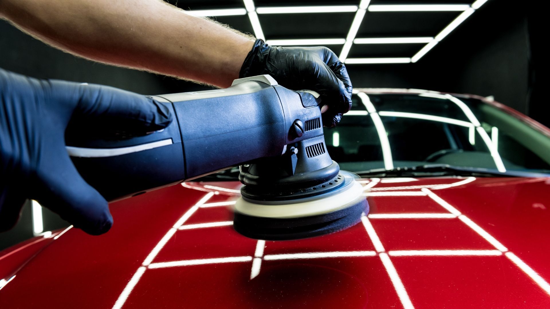 Hands polishing a red car hood with a buffer in a garage under bright lights