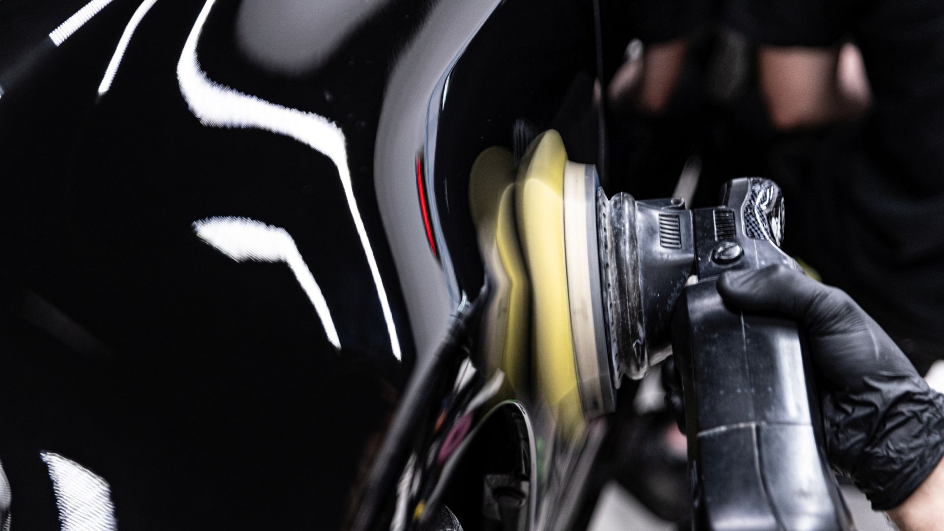 Gloved hand polishing a glossy black car with a buffing tool, creating a bright reflection