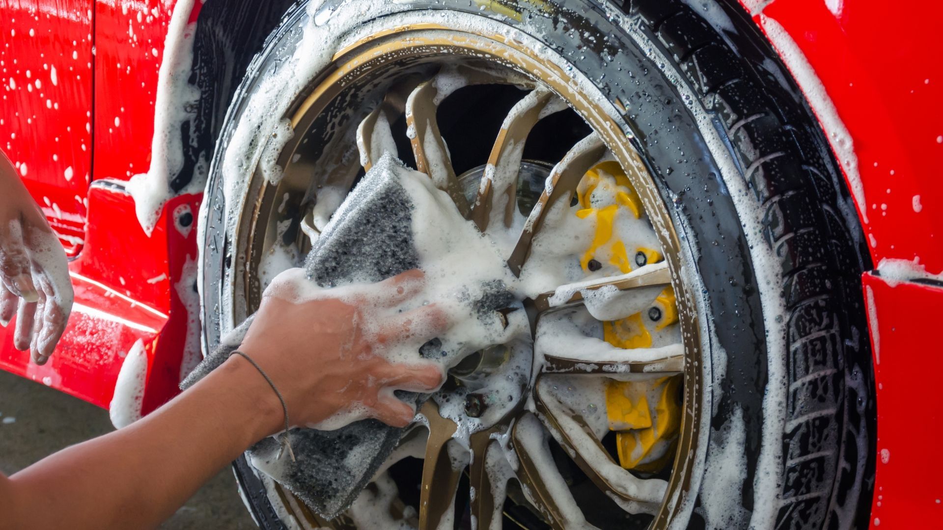 Hand washing foam off a red car’s alloy wheel and tire with a sponge