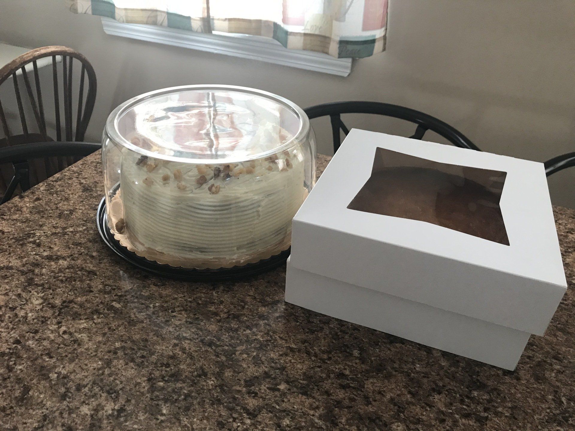 Two cakes on a kitchen counter, one covered with plastic wrap and one in a white bakery box.