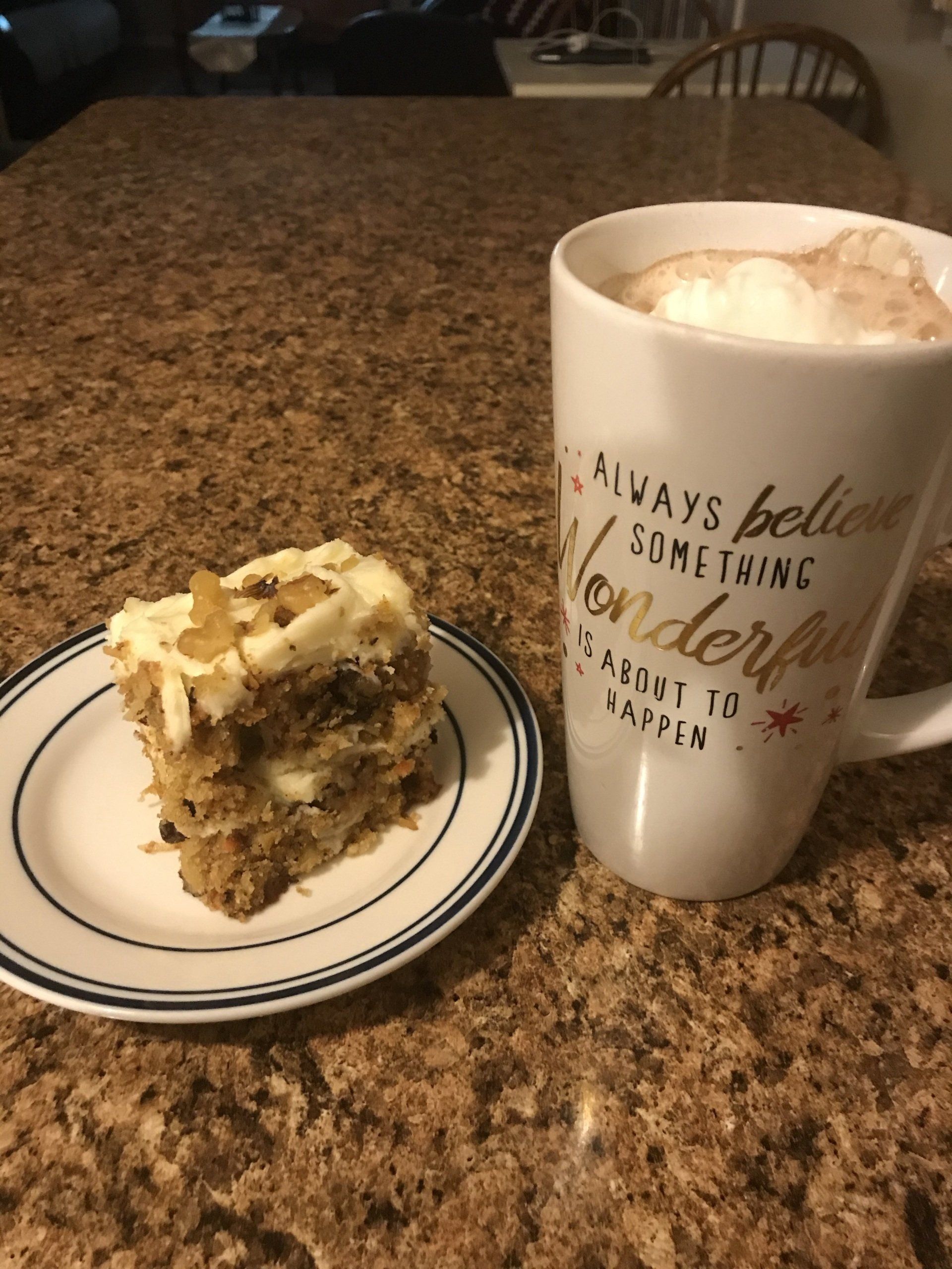 Slice of cake on a plate beside a white mug of whipped coffee on a granite countertop.