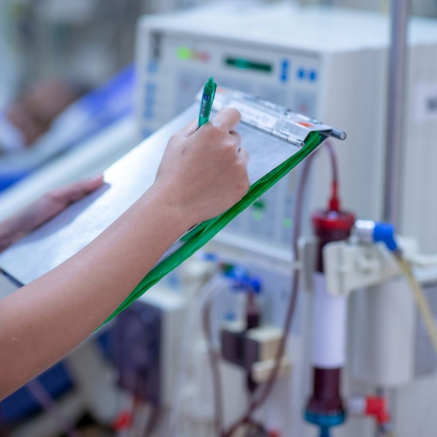 A smiling Latina nurse in navy scrubs and blue gloves stands beside a clear IV bag hanging from a stainless-steel pole. The IV bag displays a dark blue Pharmko label with the text “Intradialytic Parenteral Nutrition,” featuring a green “IDPN” badge and a medical icon. Both the nurse and the IV setup are shown clearly against a clean, neutral background.
