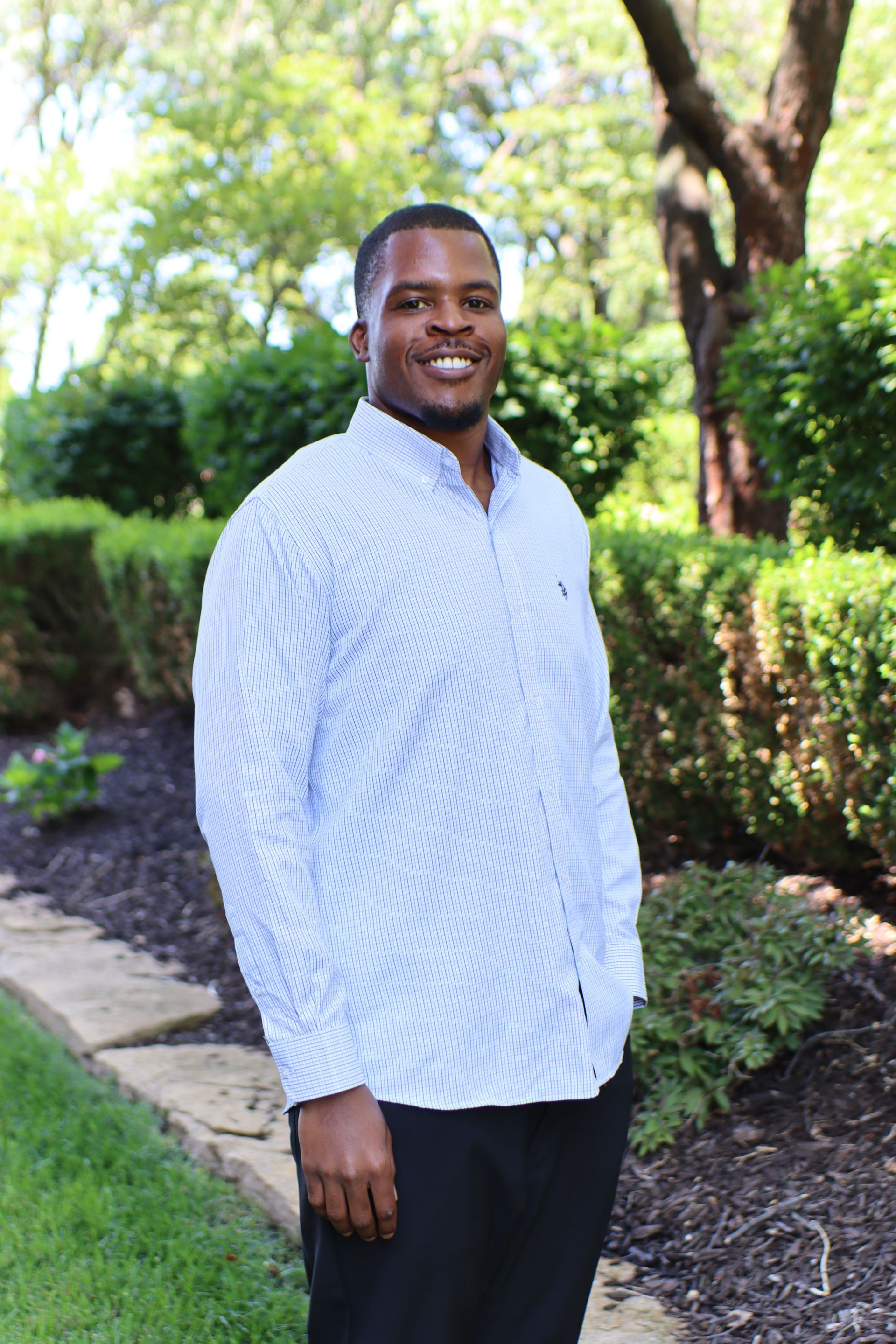 Man standing on a garden path, smiling, in a white patterned shirt and black pants.