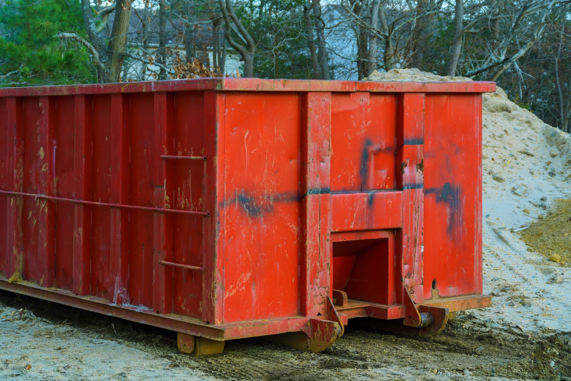 Red metal dumpster sitting on gravel beside a sandy pile in a wooded outdoor area