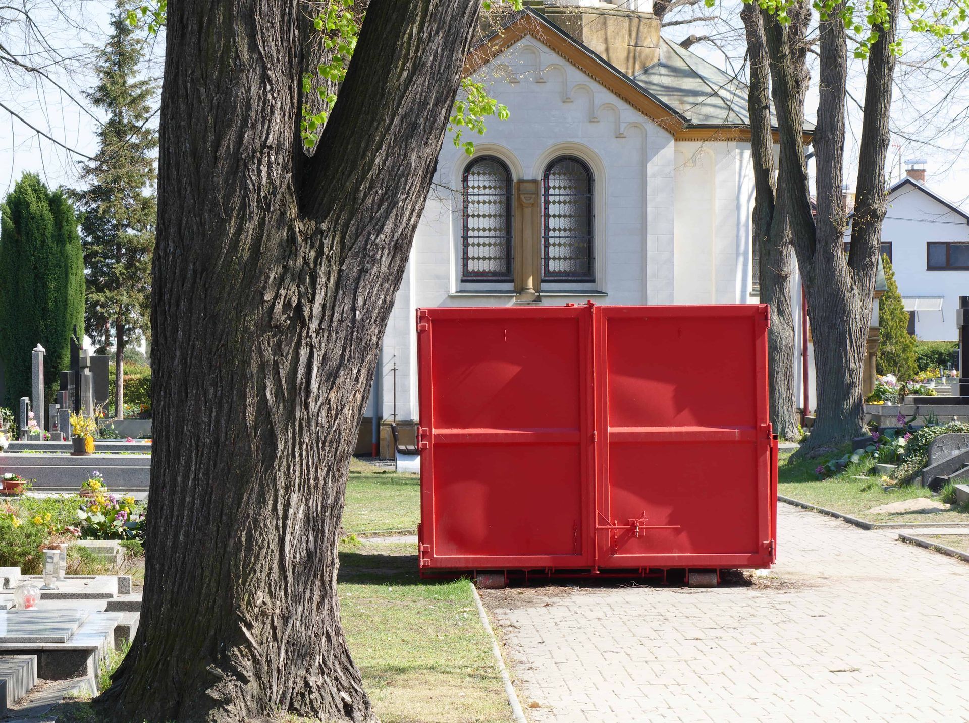 Red metal dumpster on a sidewalk beside a tree and houses in a residential area