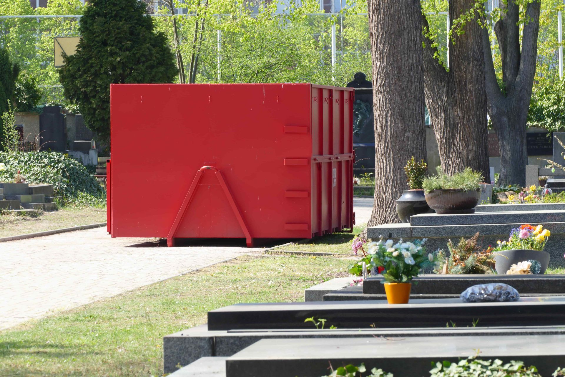 Bright red shipping container beside a tree-lined park path with benches and flower planters