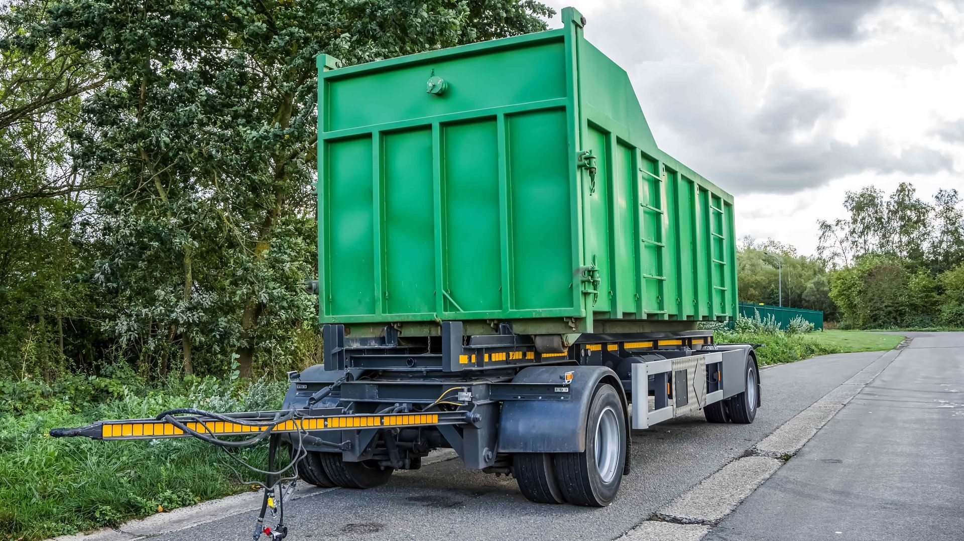 Green dumpster container on a trailer parked on a roadside beside trees and grass.