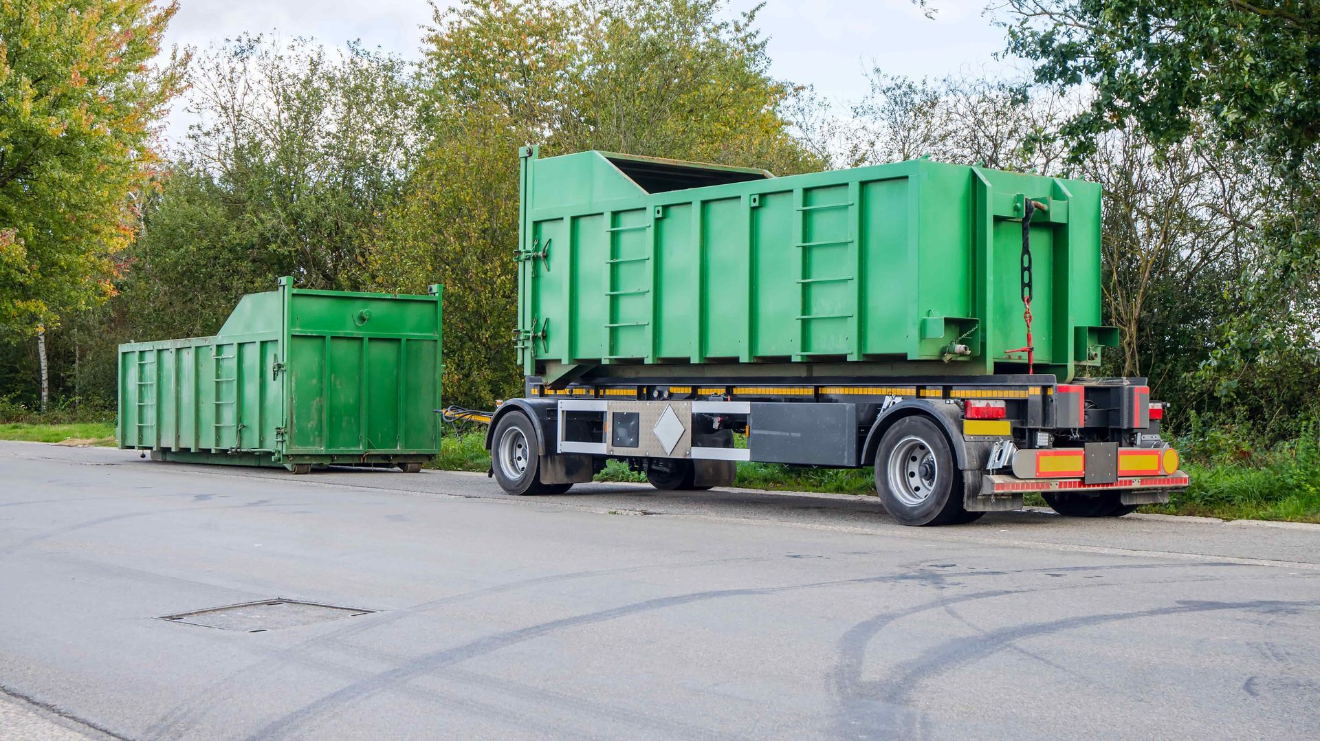 Two green industrial dumpsters on a trailer and the ground beside a paved lot with trees.