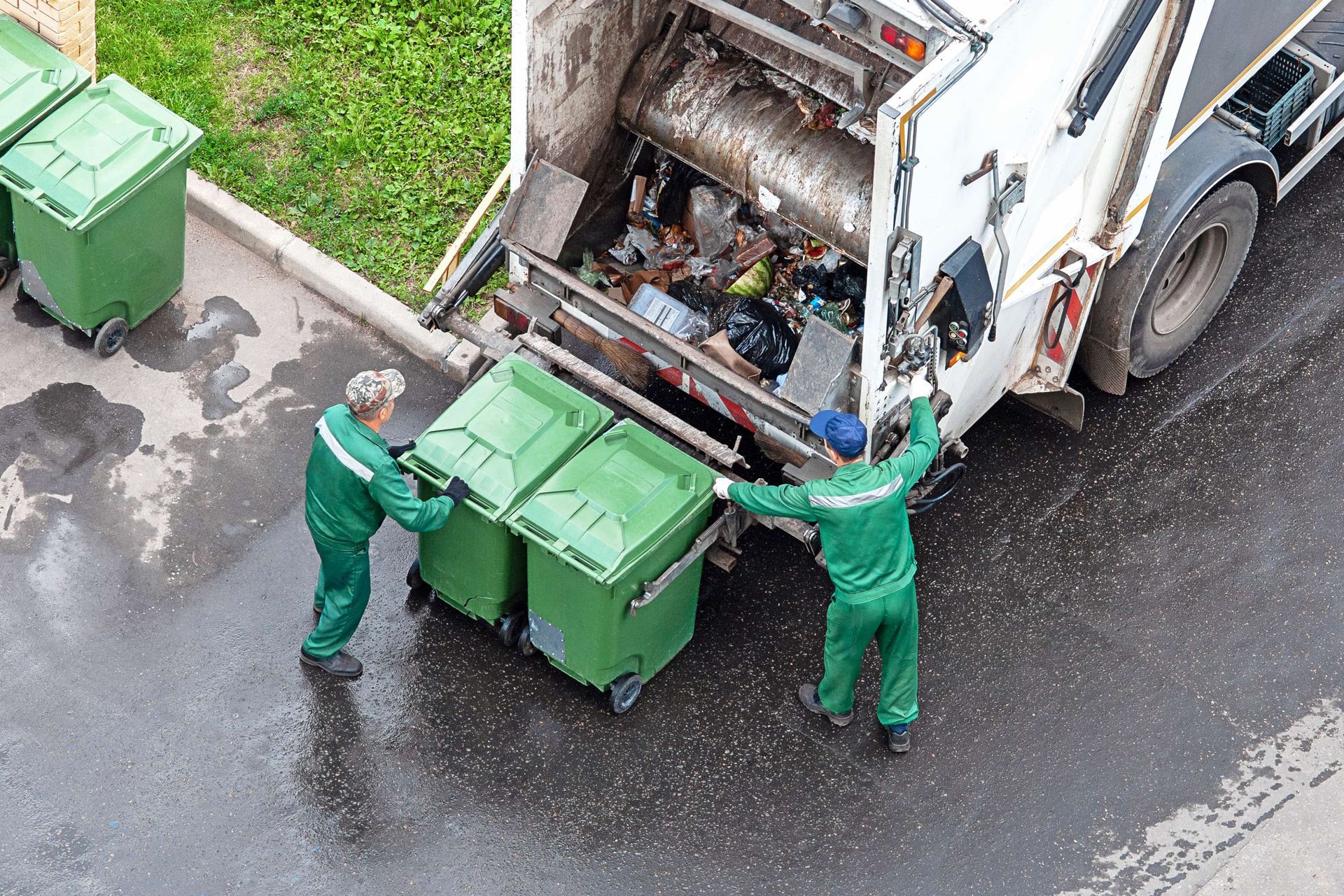Workers in green uniforms empty trash bins into a garbage truck on a wet street.