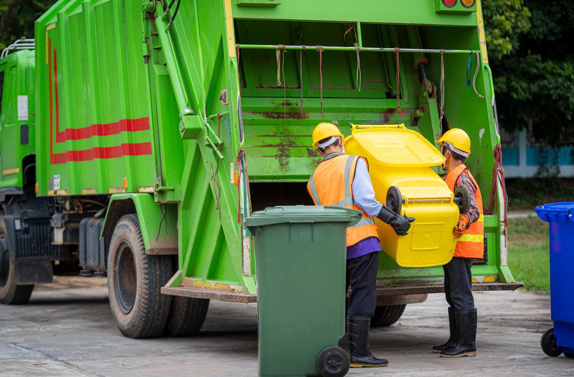 Workers empty a green trash bin into a green garbage truck on a street