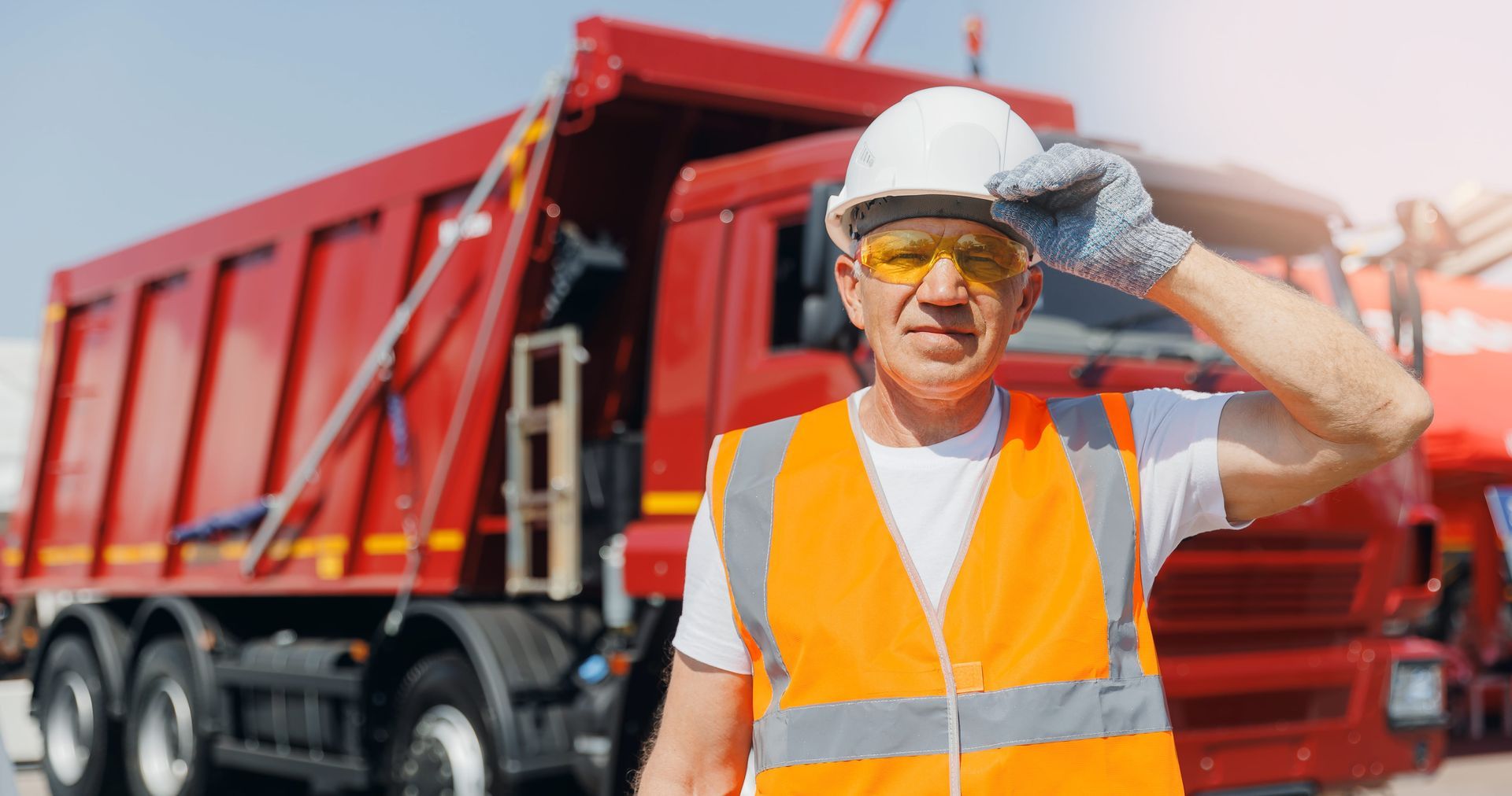 Worker in orange safety vest and hard hat standing before a red dump truck, saluting with a gloved hand.