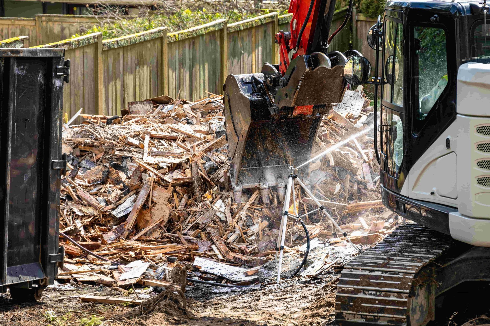 Excavator demolition bucket sorting debris in a fenced yard, with dust and rubble around it.