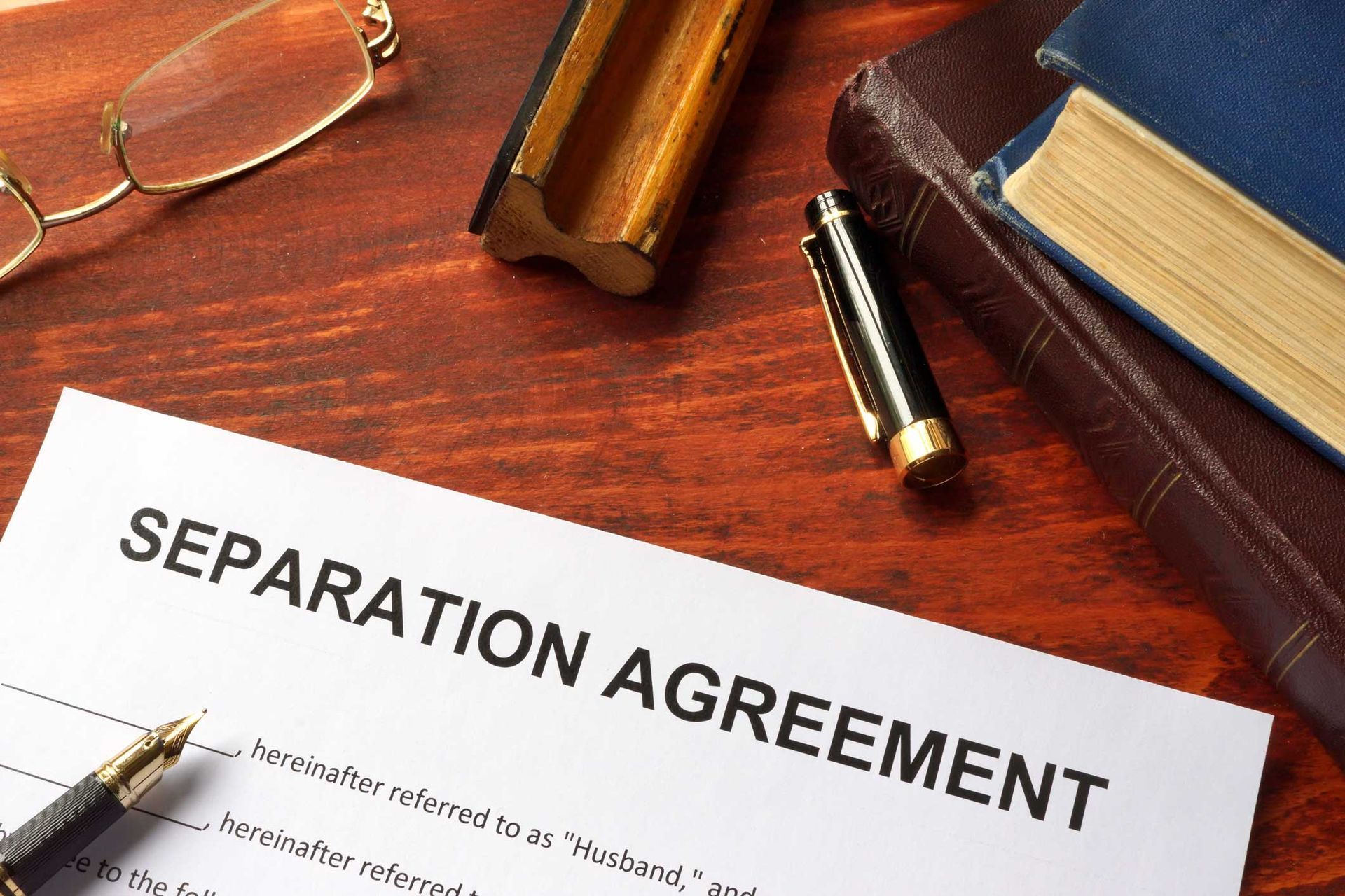A separation agreement document, pen, glasses, ink stamp, and books on a wooden desk. A separation agreement document, pen, glasses, ink stamp, and books on a wooden desk.
