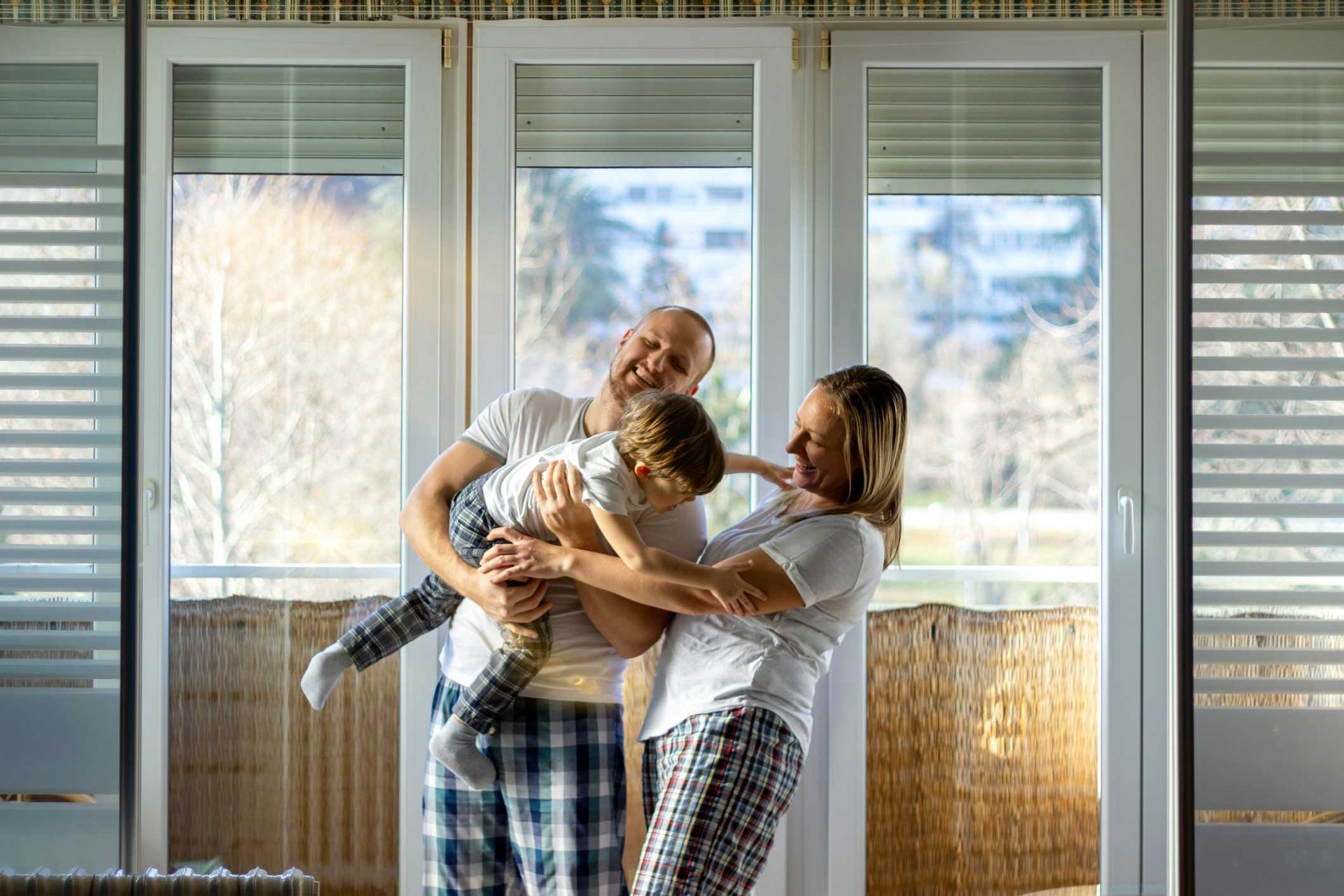 Family smiling and embracing in front of window. Child in mid-air, playing, wearing pajamas.