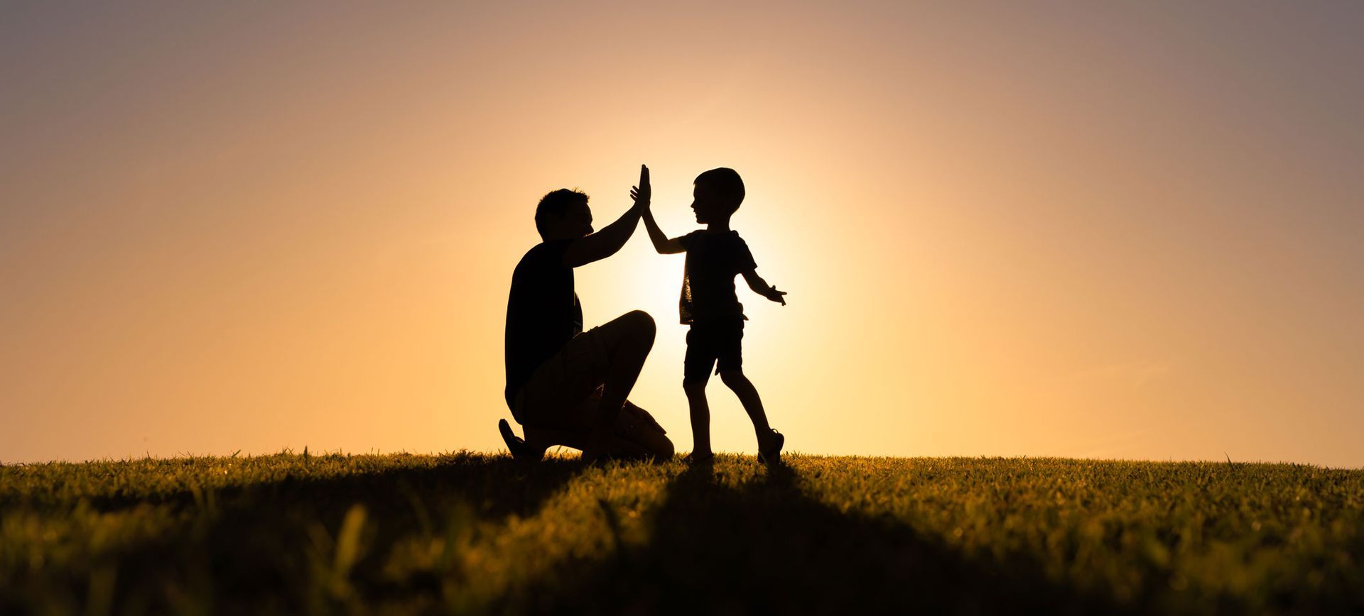 Silhouette of a person kneeling, high-fiving a child on a grassy hill with a bright sunset in the background. Silhouette of a person kneeling, high-fiving a child on a grassy hill with a bright sunset in the background.
