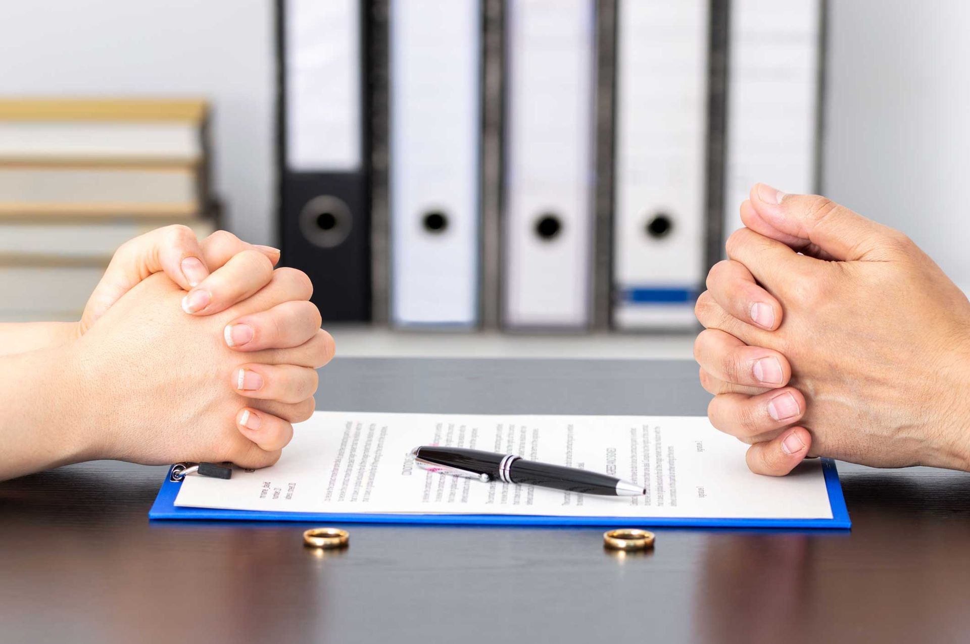 Hands clasped above a divorce agreement with wedding rings and pen, office setting. Hands clasped above a divorce agreement with wedding rings and pen, office setting.