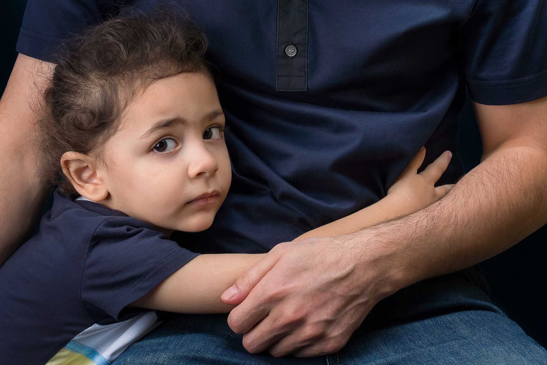 Child hugging a person wearing a navy blue shirt and jeans. The child has a worried expression. Child hugging a person wearing a navy blue shirt and jeans. The child has a worried expression.